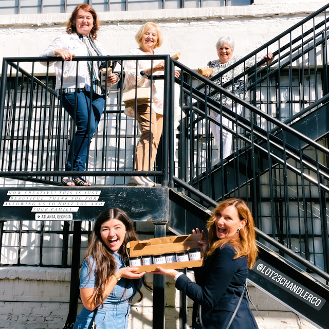 Four women posing on an outdoor black metal staircase, with one in the foreground holding a wooden box containing a set of piano keys. The background is a white brick building. At lot 23 chandler co.