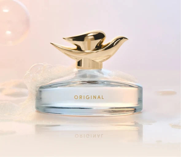 A clear glass perfume bottle with a reflective gold cap shaped like a bird, labeled 'ORIGINAL' in gold lettering, set against a soft pastel pink and white background with subtle bubbles and foam.