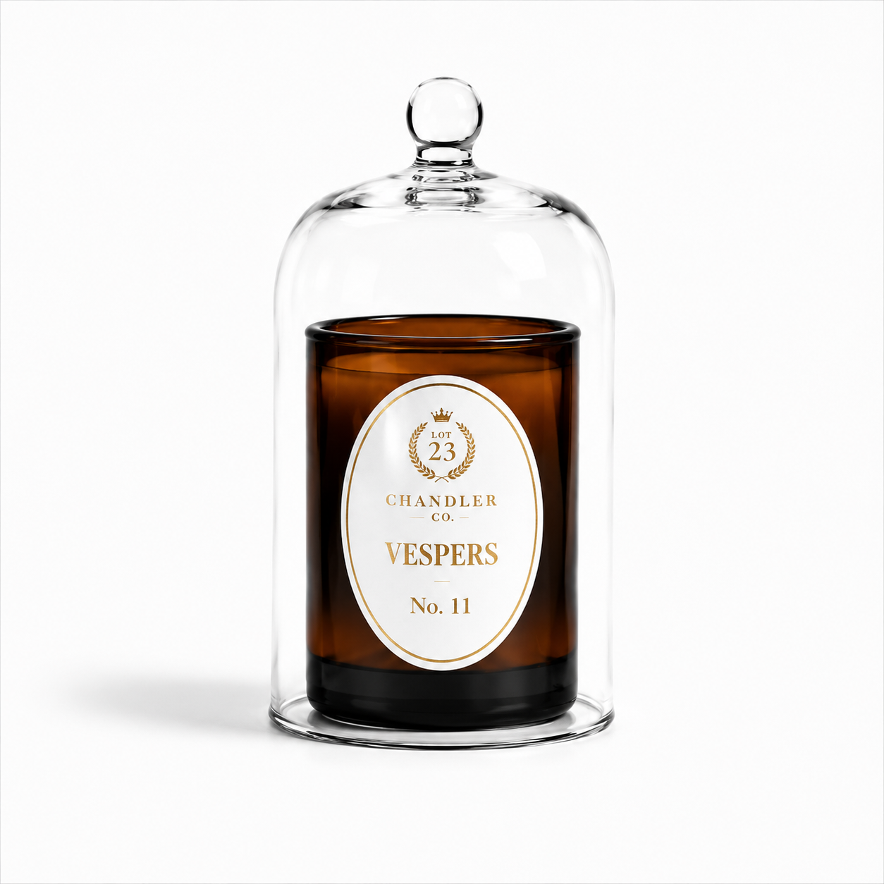An amber glass jar candle under a clear glass cloche dome with a small knob top, featuring a white oval label for Lot 23 Chandler Co. Vespers No. 11.