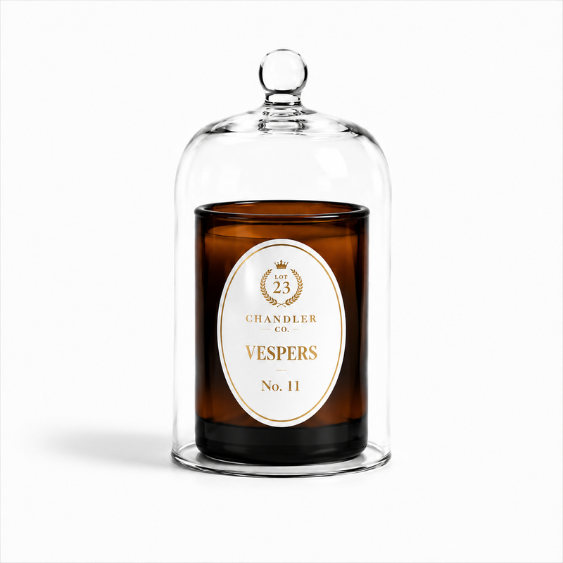 An amber glass jar candle under a clear glass cloche dome with a small knob top, featuring a white oval label for Lot 23 Chandler Co. Vespers No. 11.