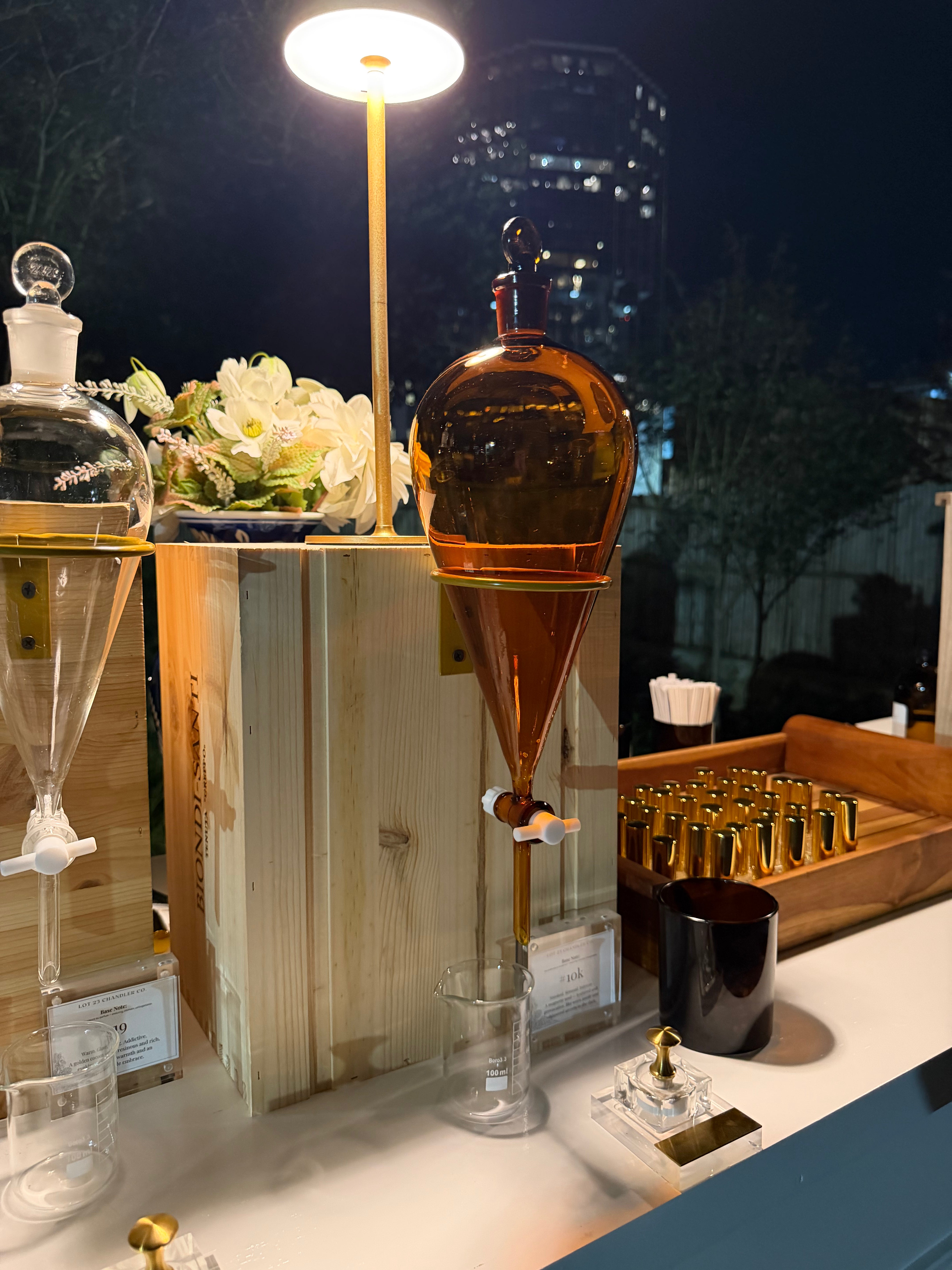A perfume bar station at night featuring a central amber glass pour-over dripper on a wooden stand, surrounded by glass perfume bottle, small gold-toned sniffers, and a lit lamp against a dark city skyline background in Atlanta.