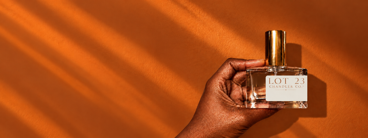 A hand holding a clear glass perfume bottle with a gold cap and a white label that reads 'LOT 23 PERFUMERY' against a warm orange background with diagonal shadows.