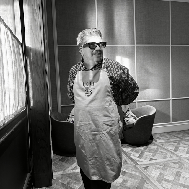 A black and white photograph of a smiling man wearing thick sunglasses, a patterned shirt, and a light apron with the text 'LOT 23' standing in a room with patterned chairs and tiled flooring.