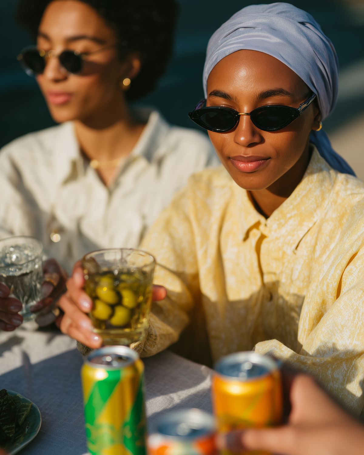Two women sitting at a table outdoors, sharing a drink. The woman in the foreground wears a light blue headwrap, black cat-eye sunglasses, and a pale yellow patterned shirt, holding a glass of iced tea with olives.